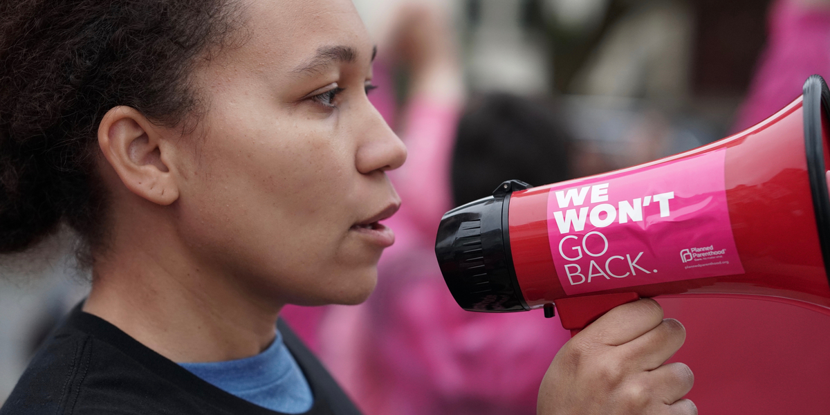 A woman with a serious expression holds a megaphone to her mouth with a sticker on it that reads WE WON'T GO BACK; behind her, a blurred crowd like a protest or audience is suggested.