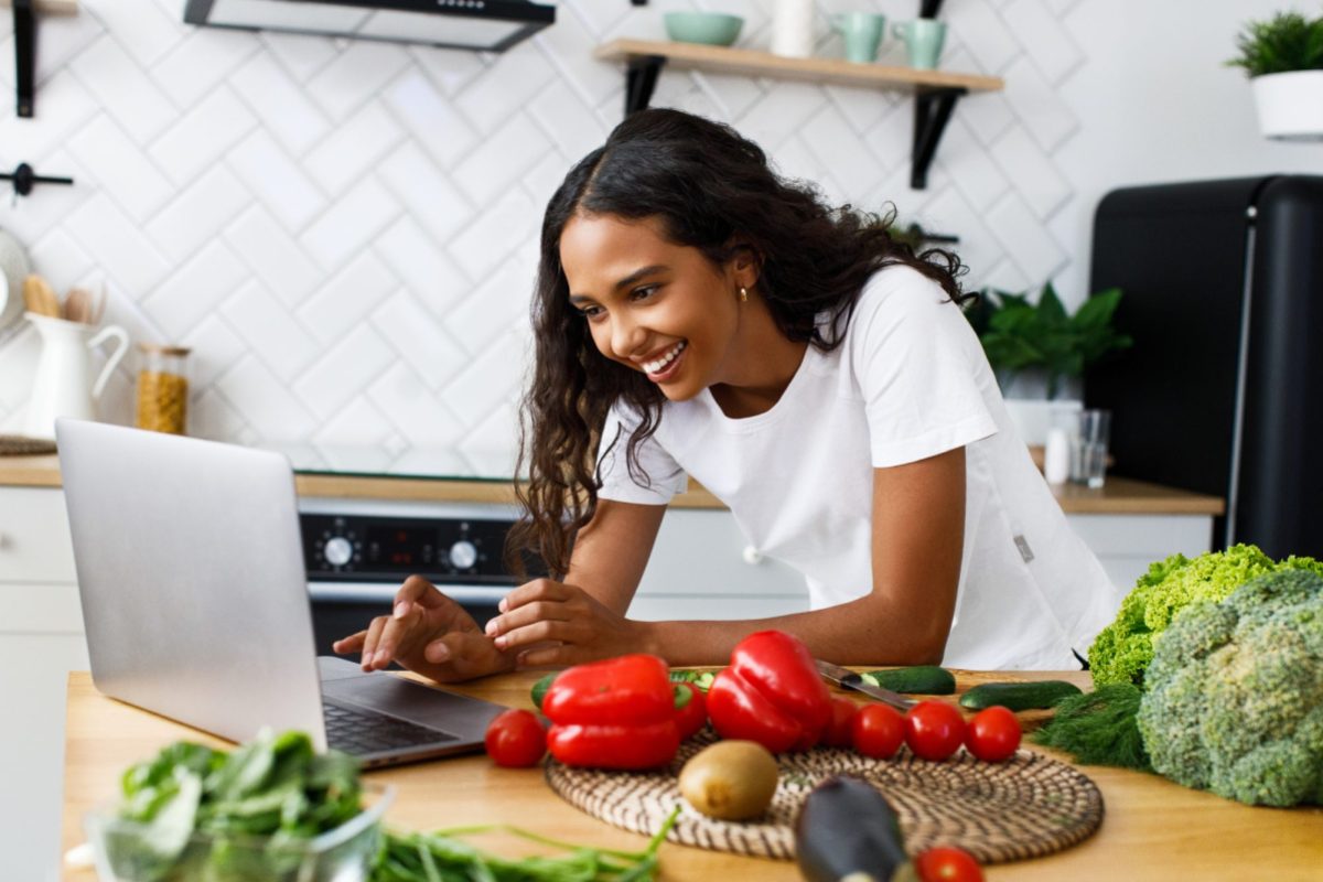 a woman leans over her computer in her kitchen. the counter has peppers, tomatoes and broccoli out and ready to cook