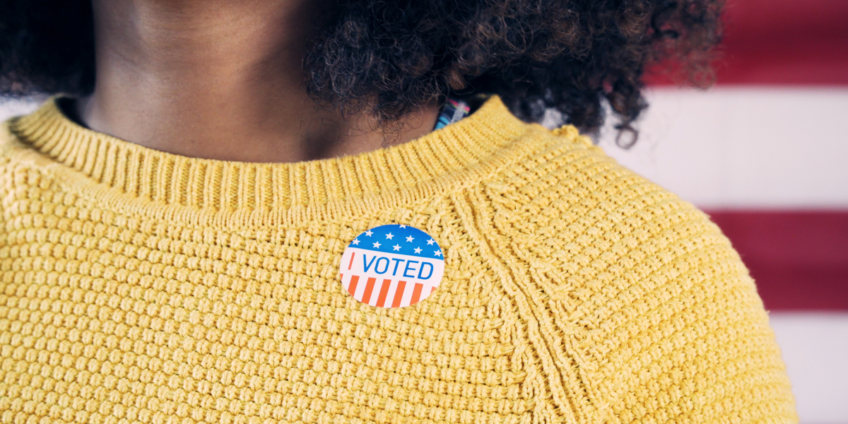 A closeup of an "I Voted" sticker on a bright yellow sweater being worn by a Black woman with shoulder-length natural hair