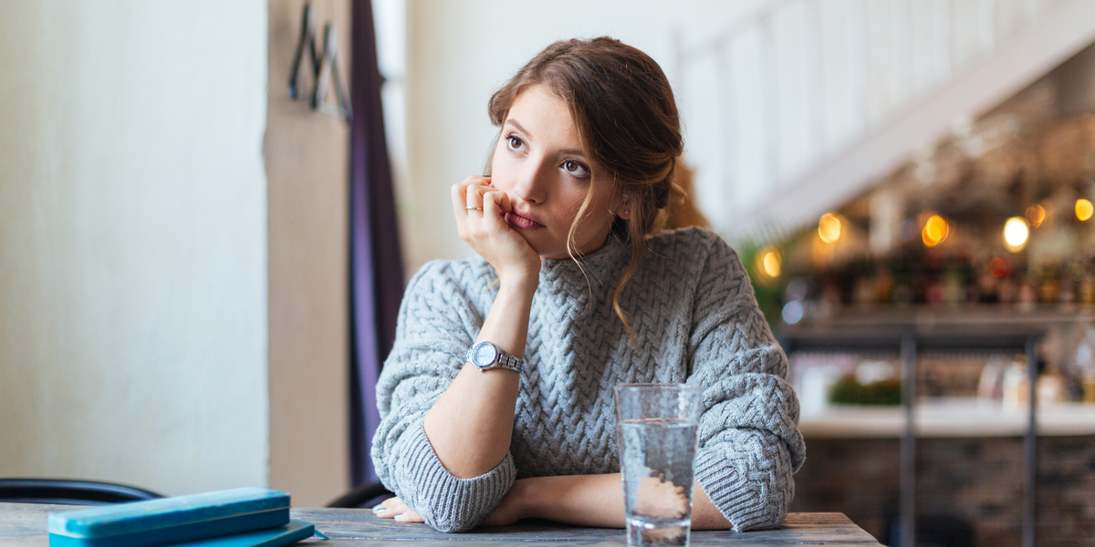 A thin white woman in a baggy sweater stares into space, looking bored, at a café table with a glass of water in front of her