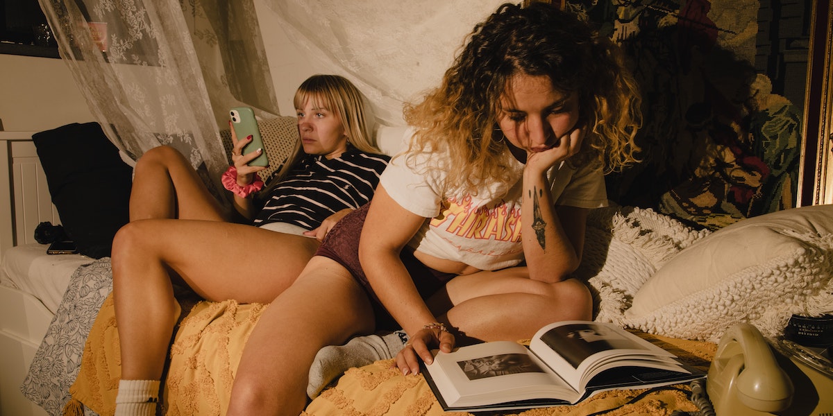 Two women are lounging in a cozy indoor space covered in blanket and pillows with a warm yellow light as if by a flashlight at night, one person reading from their phone screen and one hunched over reading from a hardback book