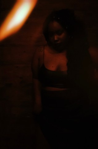 A photo of a woman in a dark room with a black shirt and a dark green skirt. The tattoo on her shoulder is visible as is the wooden wall behind her.