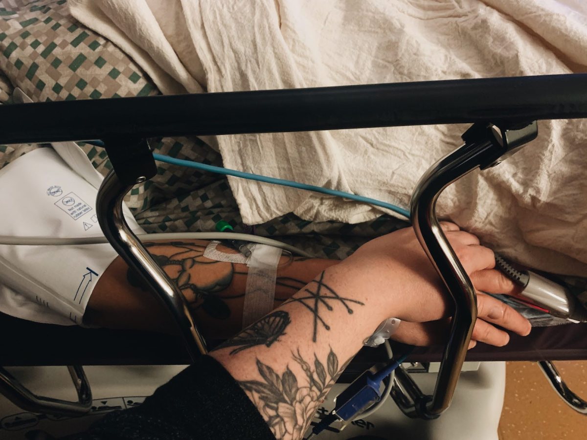 close up of two people holding hands; one woman is in a hospital bed and one is sitting next to her, both have tattoos on their forearms