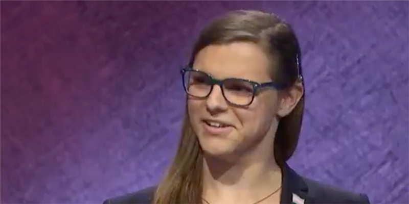 Kate Freeman, a winning contestant on Jeopardy, with thick dark blue glasses and long brown hair swept behind her ear. She is against a purple background.