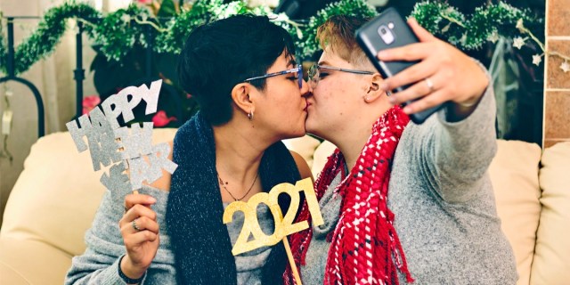 Two lesbians with short hair kiss while taking a selfie on their phone and holding up silly, sparkly "Happy New Year" signs.