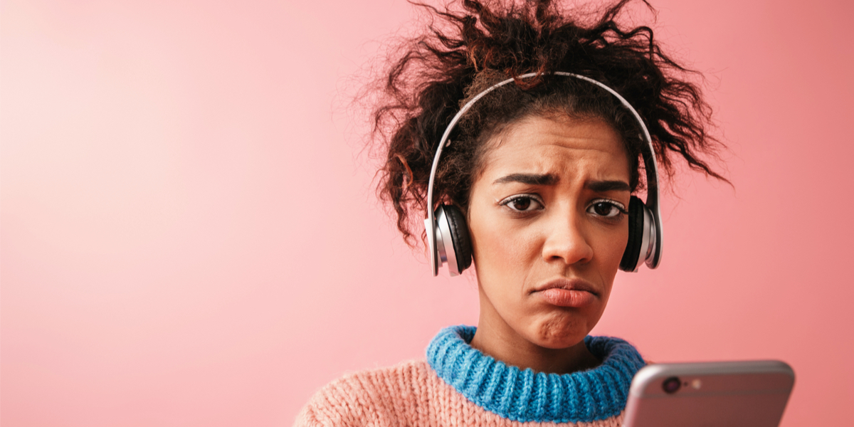 A sad lesbian listens to sad music on her sad iPhone, she's against a warm pink background.