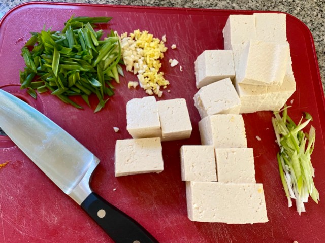 a red cutting board with green onions and blocks of tofu cut up on it