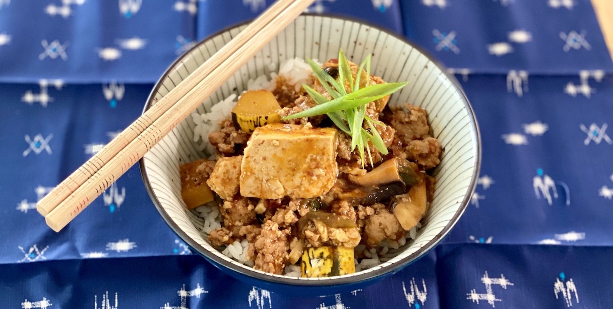 a bowl of mapo tofu over rice topped with a star of sliced scallions. chopsticks rest on the left side of the bowl and the bowl in on a blue woven cloth.