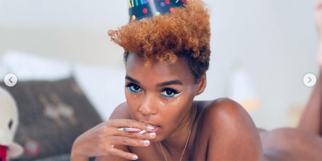 A photograph of Janelle Monáe in a birthday hat and very little else. She smiles at the camera with her eyes, her hands covering her lips, and has short red hair.