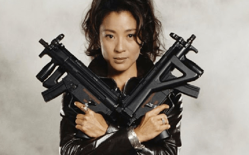 Michelle Yeoh in a leather jumpsuit holding two large guns crossed against her chest.