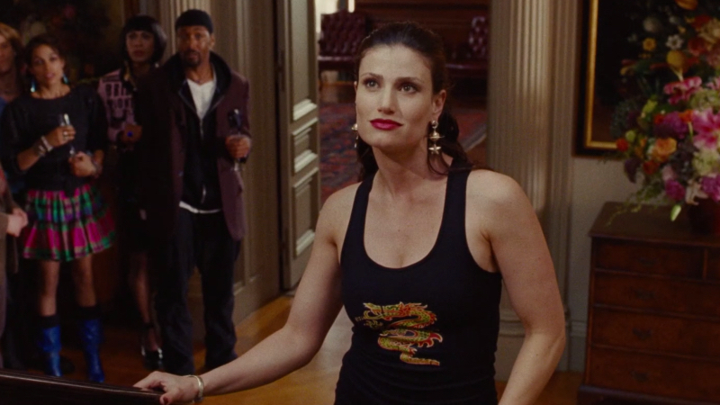 Idina Menzel stands at the bottom of the stairs in a tank top with a dragon on it and a glimmer of attitude in her eye.