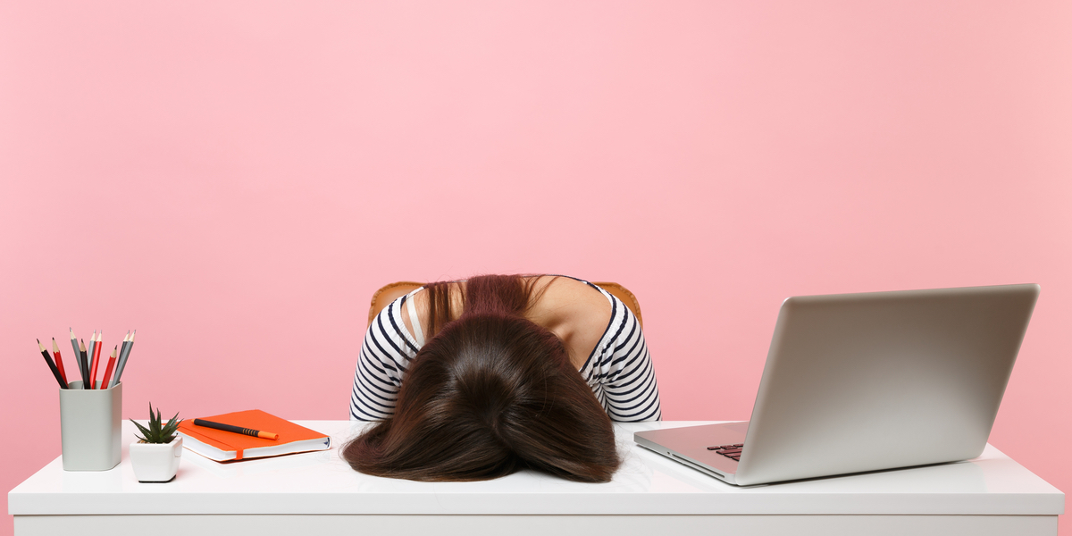 A woman with long, brown hair rests her face on her desk in front of a pink background.