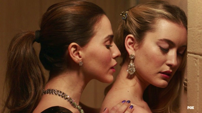 Daisy, a woman with a brunette ponytail, kisses Becky, a blonde woman with her hair swept to the side, at the lower part of her neck. It's small and intimate.