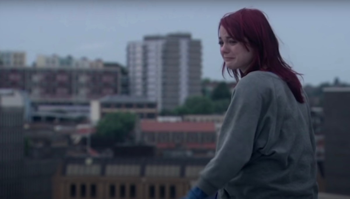 A scene from Skins where Emily Fitch sits on a concrete roof, looking upset with the Bristol skyline behind her