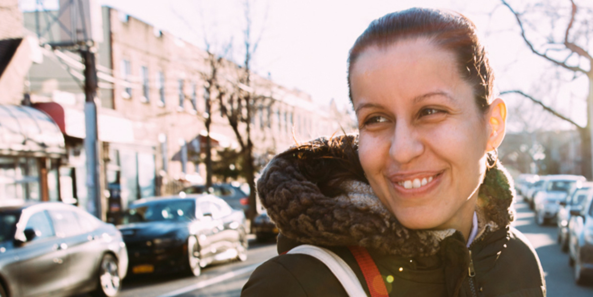 A photo of Tiffany Cabán, a queer latina with a long pony-tail, looks over her shoulder and her parka hood as she crosses the street.