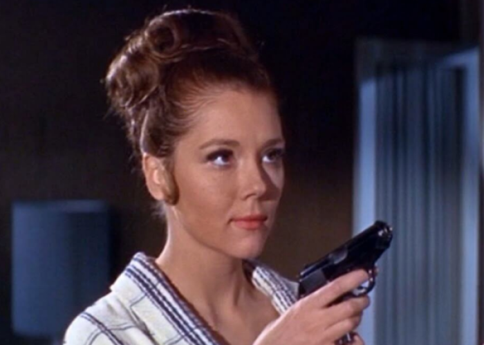 A brunette in a 1960s style updo holds a small pistol while wearing a silk robe.