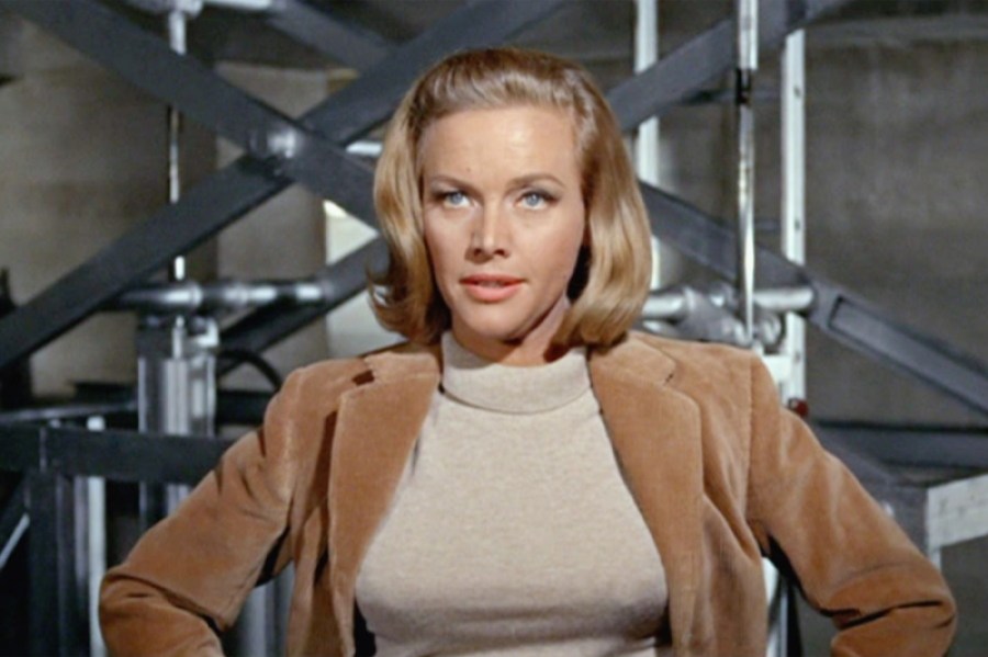 A blonde woman with a bob haircut stands with her hands on hips in a camel colored blazer.