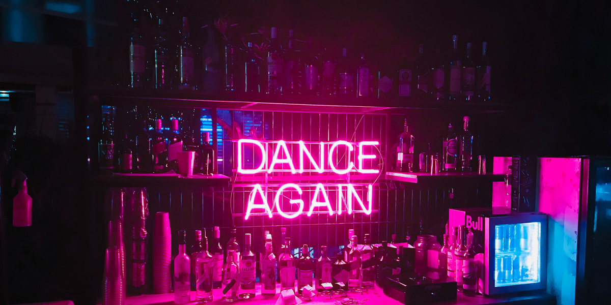 A neon pink sign says "Dance Again" against a bar wall with empty bottle everywhere.
