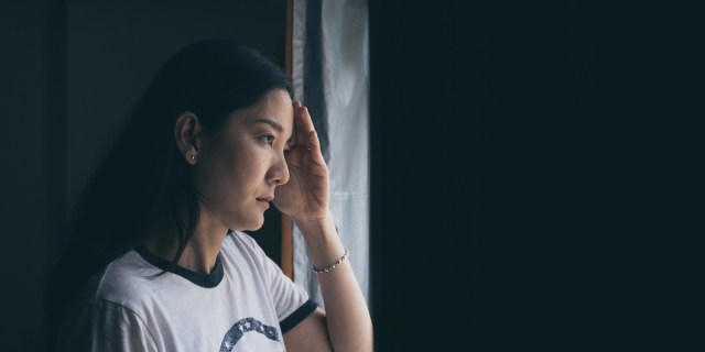 A woman in a white T-shirt with long, black hair holds her hand to her forehead and looks out the window.
