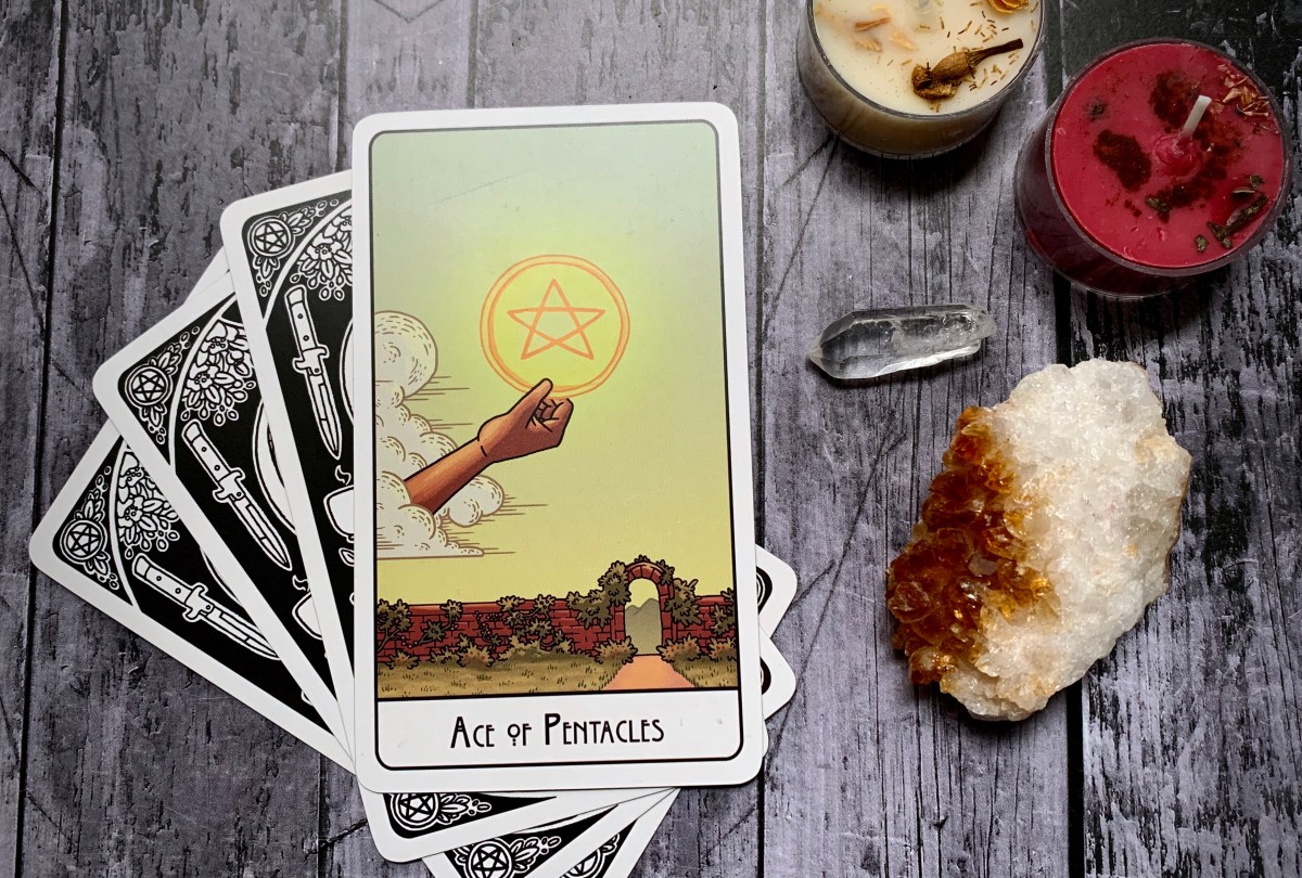 The Ace of Pentacles card sits on top of three other face-down cards. The Ace depicts a disembodied hand holding a glowing pentacle against a clear sky, below is a stone wall with a gate in it.