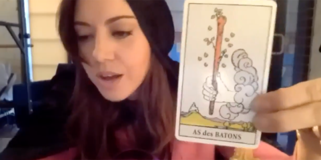 Aubrey Plaza holds up a tarot card while wearing a black cape.