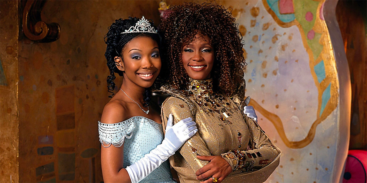 Still from Roger and Hammersteins 1997 "Cinderella". shows the two stars, Brandy and Whitney Houston, Brandy is wearing a pal blue ball gown and tiara and Whitney Houston is wearing a gold gown.