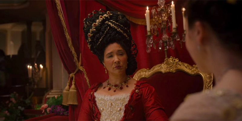 Image shows The Queen in a red and cream gown sitting in a chair having conversation. Her hair is in box braids and layered on top of each other into a high stacked style. It's adorned with gold and red ribbons and more, and topped with a crown.