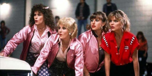 Still from Grease 2. 4 girls wearing Pink Satin Jackets and one in a red bowling shirt, all lean atop a car.