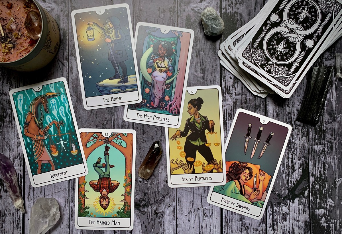 There are six cards laid out in this photograph. Judgement depicts what appears to be the Egyptian god Anubis, holding a scale. On one end of the scale a human heart, on the other, a feather. In the background are a number of souls ascending against a teal backdrop. The Hermit card depicts a lone femme person with long silver or gray hair, in contemporary winter clothing, standing on the edge of a snowy cliff. They hold up a lantern and have a walking stick in hand. The Hanged Man card depicts a mas person in lumberjack-esque clothing, hanging upside down by their foot, from a tree. Their knee crosses, forming a triangle, and a bird rests atop their shin. The High Priestess card depicts a femme person, seated on a crescent moon, a glowing book floats between their hands. To the left is a white tree, and to the right is a dark brown tree. The Six of Pentacles card depicts a person in a business suit sprinkling gold coins to waiting hands below. Finally, the four of swords depicts a person asleep in bed, clutching one of the swords. The other three swords hang on the wall above them.