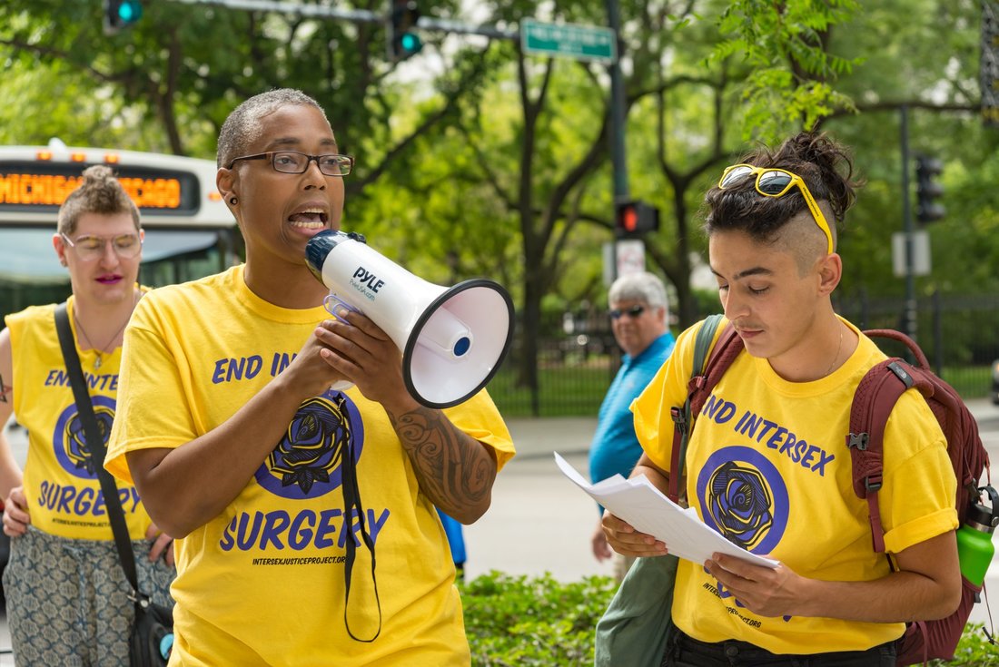 Sean Saifa Wall, who has dark brown skin and hair shaved close to the scalp, speaks into a bullhorn and stands beside Pidgeon Pagonis, who has light brown skin and the sides of their hair shaved. Pidgeon is reading a piece of paper.