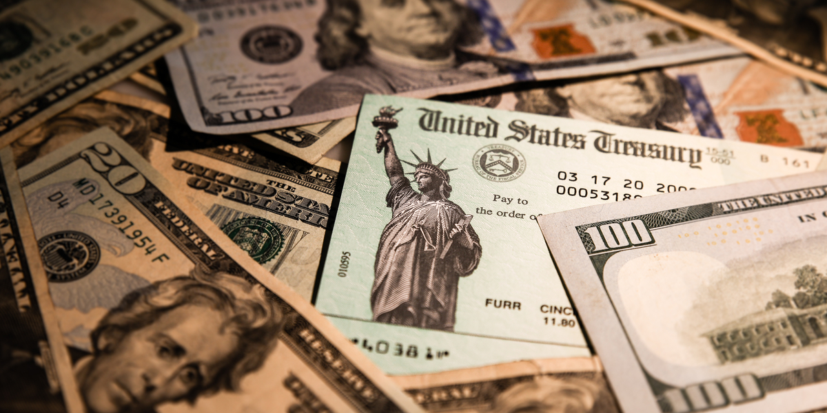 A close-up shot in dramatic lighting of a US-treasury-branded check mixed in among a pile of fresh bills in US currency.