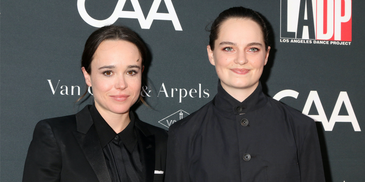 Elliott Page and their partner Emma on a red carpet event, both have their hair slicked back into low bungs and have on black suits with black collared shirts underneath.