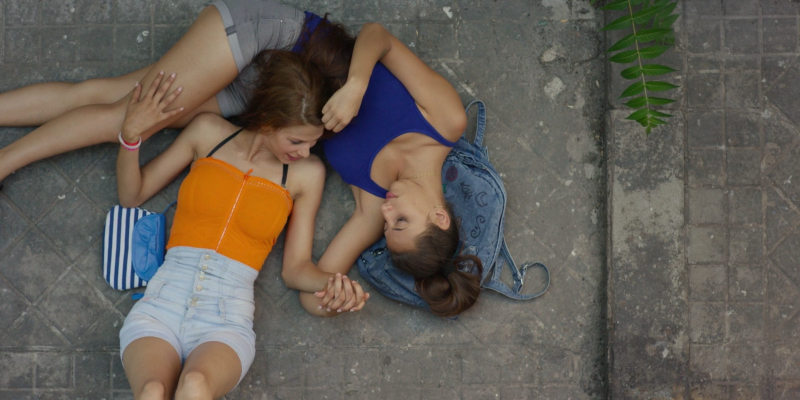 Two young women embrace on concrete ground.