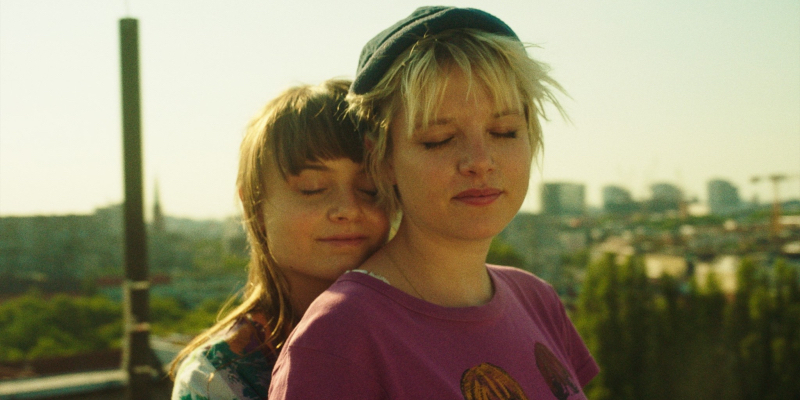 A young queer wraps her arms around another young queer with a cityscape behind her.