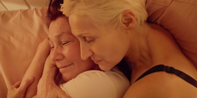 Two older women cuddle in bed.