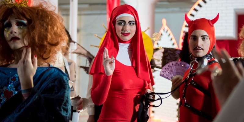 A person in a red nun costume holds their partner in a red devil costume on a leash.