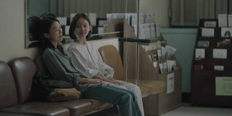  A woman sits next to a girl in a hospital gown and a bandage on the side of her head.