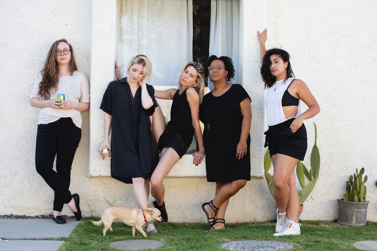 The Autostraddle Senior Staff in black and white outfits against a white wall in a California sunset. From L to R: Rachel, Laneia, Carol the dog, Riese, Carmen, and Sarah