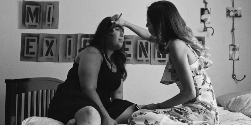 A teen girl does the makeup of another teen girl with spray painted letters on the wall behind them.