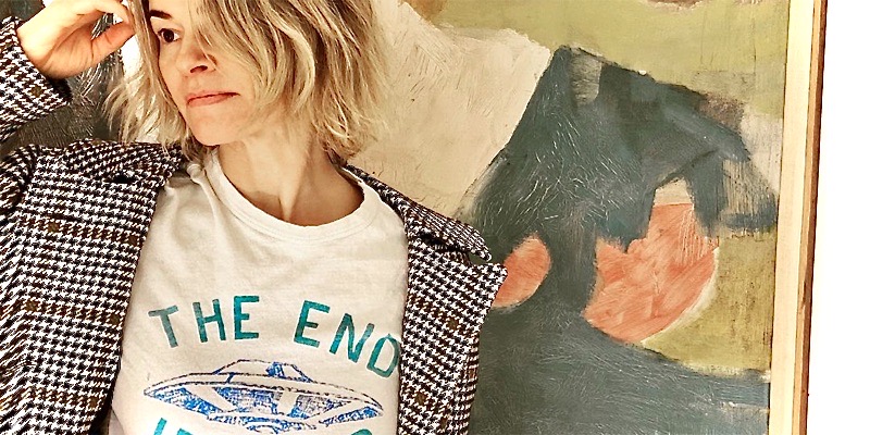 Leisha Hailey in a t-shirt that says "the end is near" with an alien spaceship in blue. She is standing next to a piece of green and yellow art.