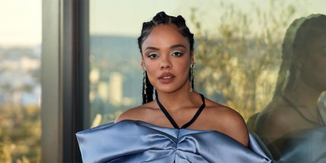 Tessa Thompson in cornrows with blue eyeshadow and a blow satin sleeveless dress stands against a mirrored window at sunset.