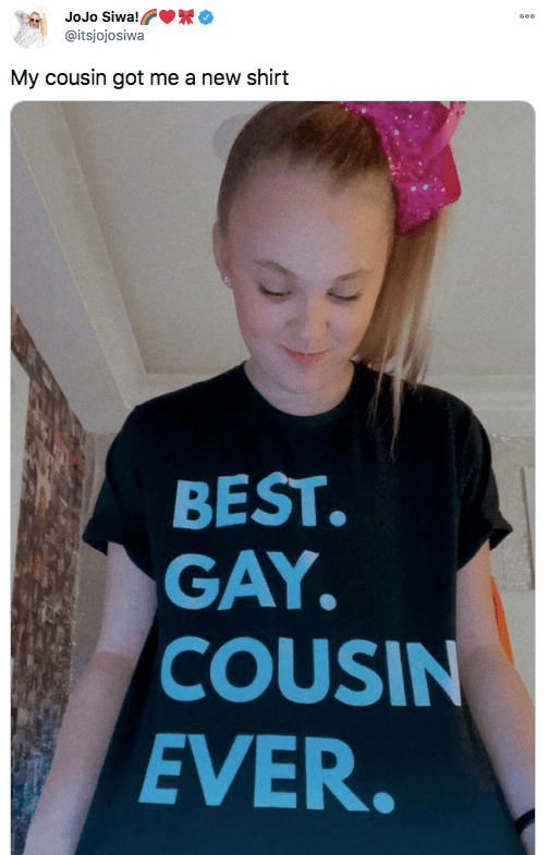 JoJo Siwa wears a pink hair bow and black shirt with blue lettering that reads, "Best. Gay. Cousin. Ever."