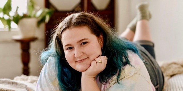 Sophie Carter-Kahn, the host of the podcast "She's All Fat" lays down on her bed with her feet crossed in the air behind her. She has dyed blue tips on her shoulder length brown hair and her face is resting gently on her hand.