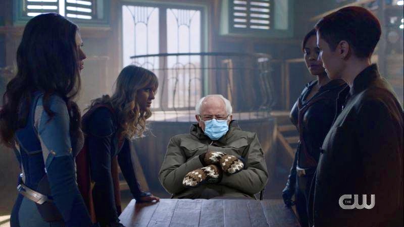 Cut out Bernie Sanders sits at a wooden table while everyone plots how to take down a villain on Supergirl