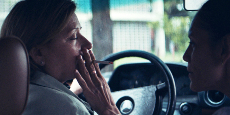A woman gives an older woman a puff from her cigarette while sitting together in a car.
