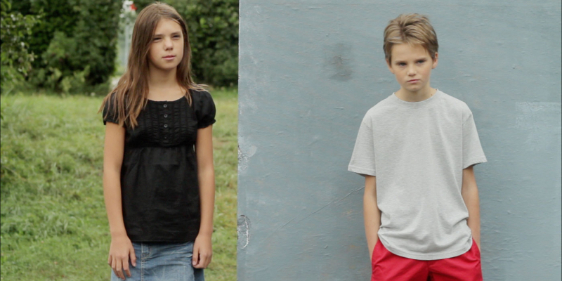 A young person dressed as a boy stands against a pale blue wall next to a girl standing in front of a grassy background.