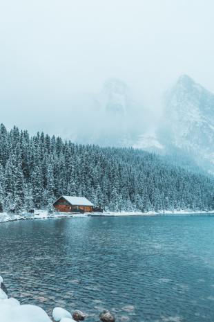 Image shows a Cabin far off in the woods. Mountains are in the background and it is sitting right off a lake - snow covers everything