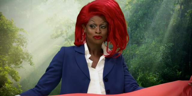 Symone from RuPaul's drag race smirks in a navy blue suit jacket with a white collared shirt and a bright red wig that is styled to the side.