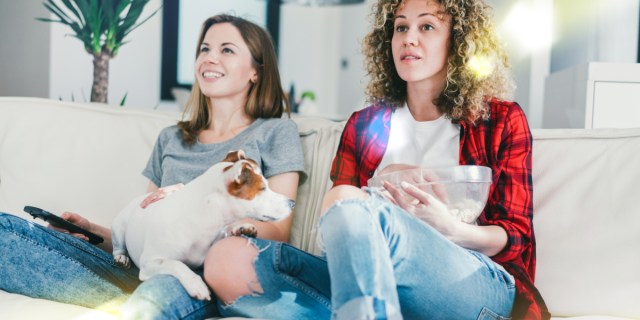 Two white women wearing a gray tshirt and red flannel respectively lounging on a couch, one smiling and one looking more serious, with a Jack Russel Terrier sitting between them, looking disgruntled
