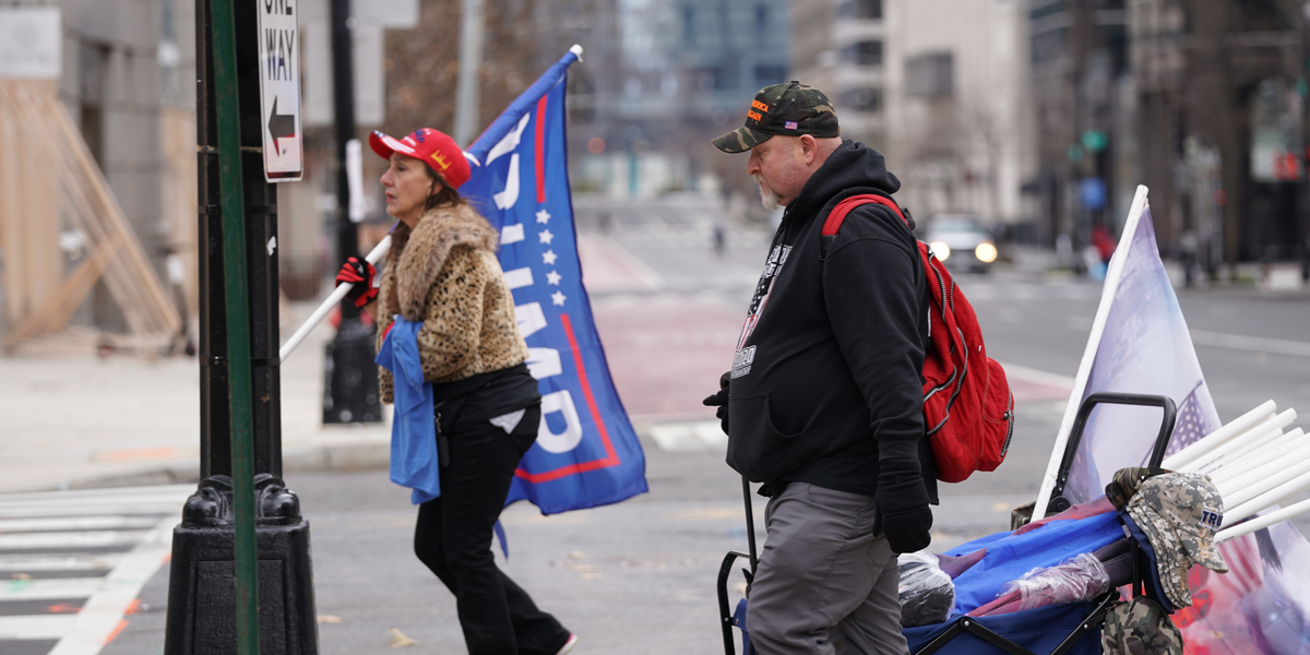 Two white people carry Trump flags and merchandise in their hands and a rolling wagon while wearing MAGA hats in DC on the morning of January 6, 2020.